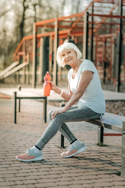 Go for sports. Old sporty smiling woman with short grey hair holding a bottle of water and sitting at the sports ground bench.