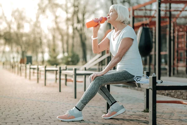 İçme suyu. Kısa beyaz saçlı, atletik giysiler giyen ve oyun alanında şişeden içme suyu ile güzel bir yaşlı spor odaklı kadın