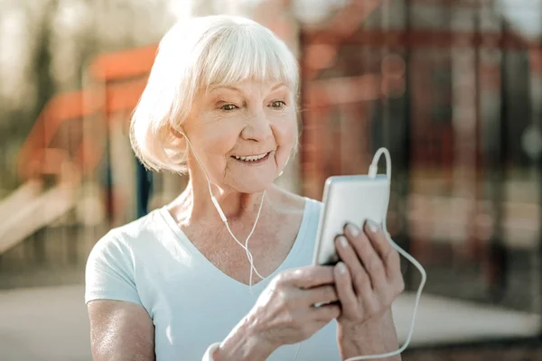 電話を持ってる白い携帯電話を持ち 集中して見ているヘッドフォンを持つ高齢者のスポーティな灰色の髪のビーミングマダム — ストック写真