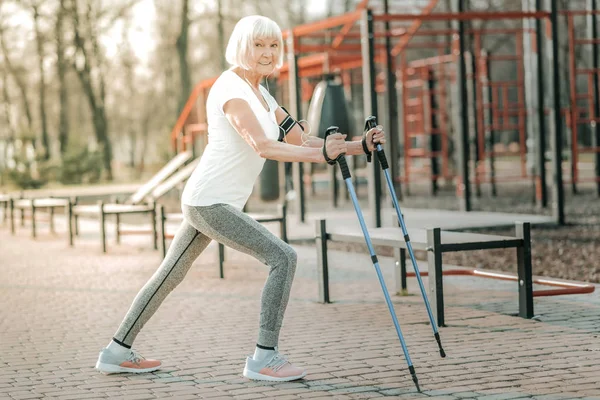 Long steps with walking poles. Appealing sporty madam in advanced years wearing workout clothing doing long steps with walking poles on the athletic ground