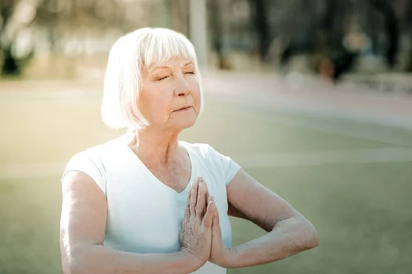 Breathing techniques. Peaceful good-looking aged madam with short silver haircut practicing breathing techniques on the green grass play yard.