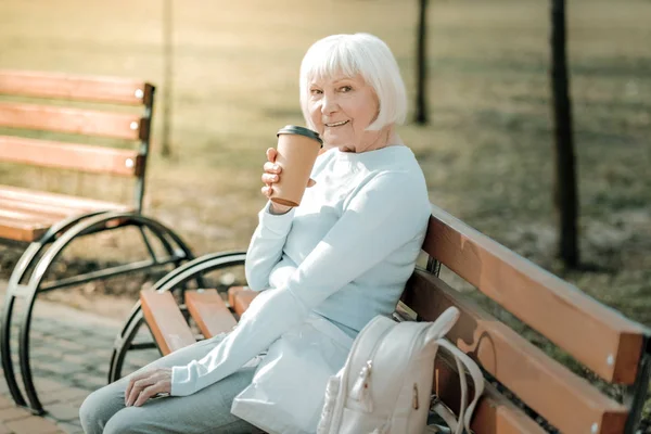 Drinking coffee to go. Well-groomed short-haired smiling senior woman drinking coffee to go in the park