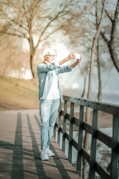 Joyful stylish elderly woman taking photo standing on the bridge — Stock Photo, Image