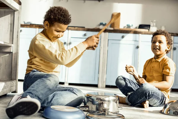 Chico balanceando cuchara para golpear la cacerola mientras su hermano está sentado cerca . — Foto de Stock