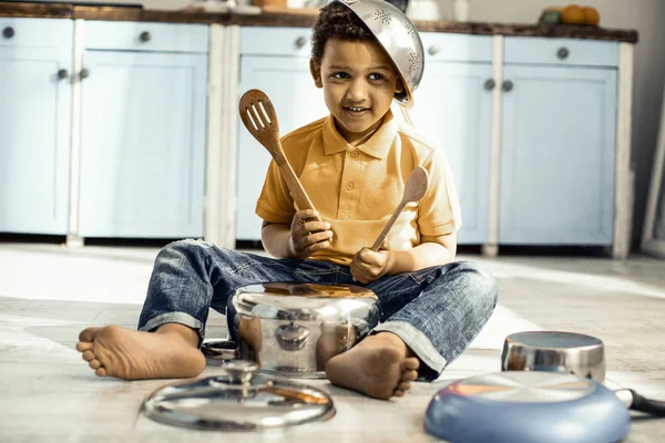 Boy sitting on the kitchen floor and playing on a saucepan while wearing a colander on the head. — Stock Photo, Image