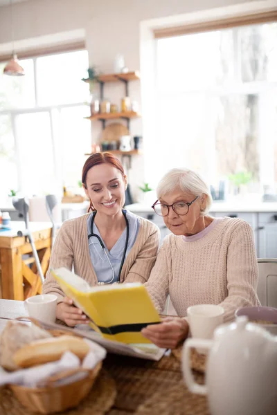 Nurse sitting near pensioner reading book in the morning — Stock Photo, Image