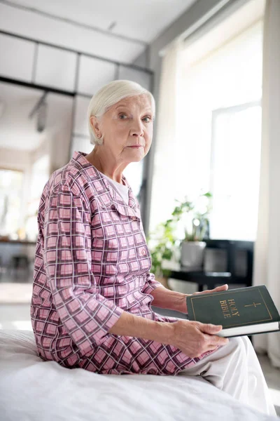 Aged lady holding Bible while sitting on sofa — Stock Photo, Image