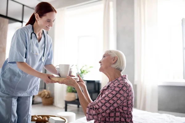 Κοκκινομάλλα νοσοκόμα φέρνοντας πρωινό για συνταξιούχος γυναίκα — Φωτογραφία Αρχείου
