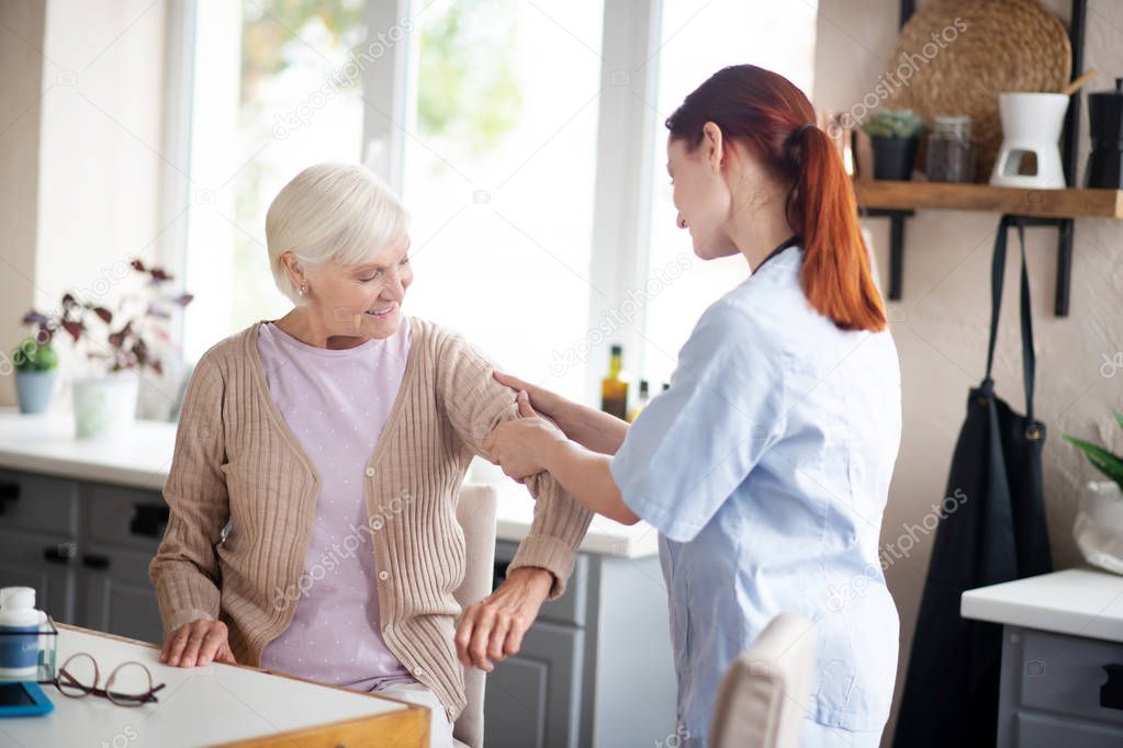 Red-haired caregiver massaging arms for retired woman