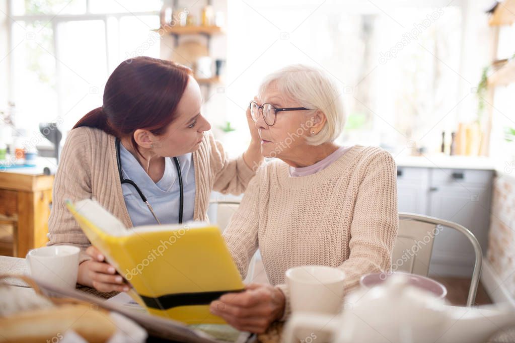 Caring red-haired nurse fixing glasses for retired woman