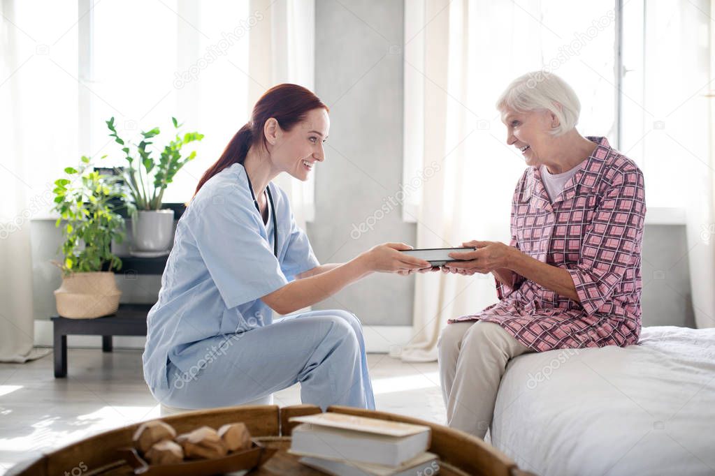 Caring nurse smiling while giving book to aged woman