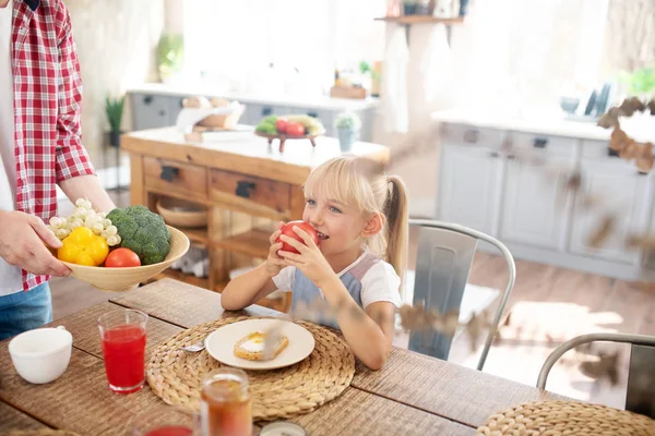 Linda hija rubia mordiendo tomate mientras desayuna —  Fotos de Stock
