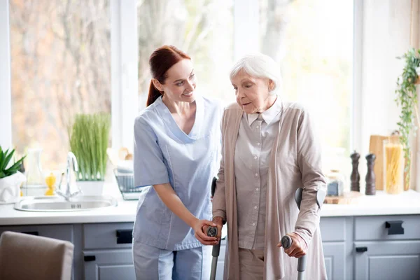 Smiling caregiver helping aged woman to make steps — 스톡 사진