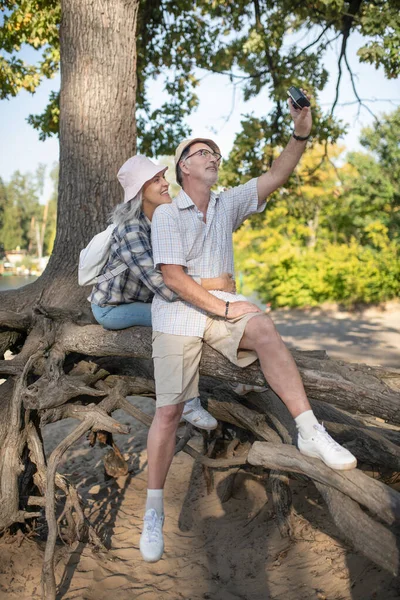 Esposa sonriendo mientras haciendo selfie con el marido sentado cerca de árbol enorme —  Fotos de Stock