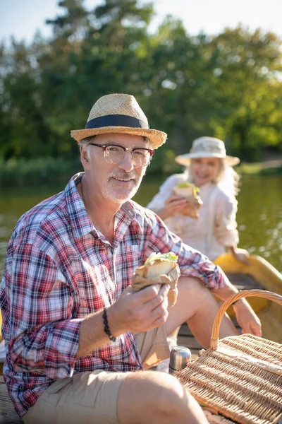 Husband wearing glasses enjoying yummy sandwich while having boat trip — Stock Photo, Image