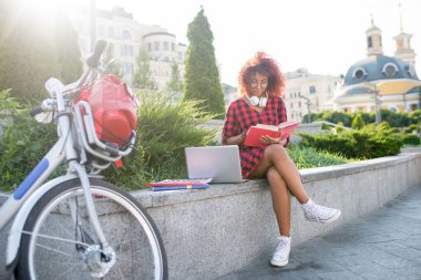 Kızıl saçlı öğrenci dışarıda otururken kitap okuyor.
