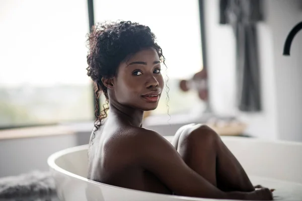 Dunkeläugige lockige Frau, die zu Hause in der Badewanne sitzt — Stockfoto