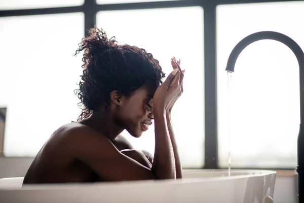 Кучерява усміхнена жінка миє обличчя під час ванни — стокове фото