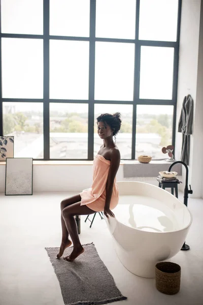 Frau in Handtuch gehüllt in der Nähe der Badewanne stehend am Fenster — Stockfoto