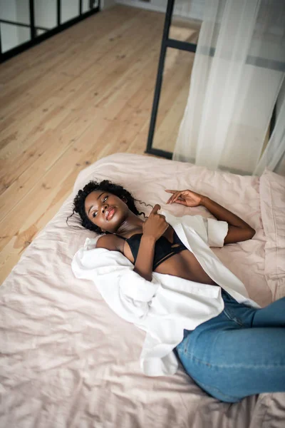 Slanke jonge vrouw dragen zwart beha en wit shirt chilling op bed — Stockfoto