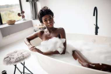 Güzel siyah gözlü kadın köpükle banyoda yatıyor.