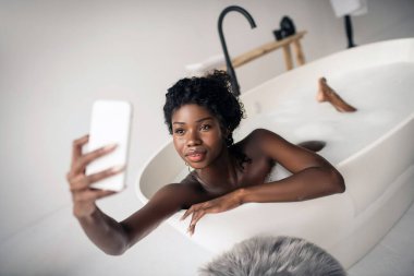 Siyah gözlü kadın köpüklü banyo yaparken selfie çekiyor.