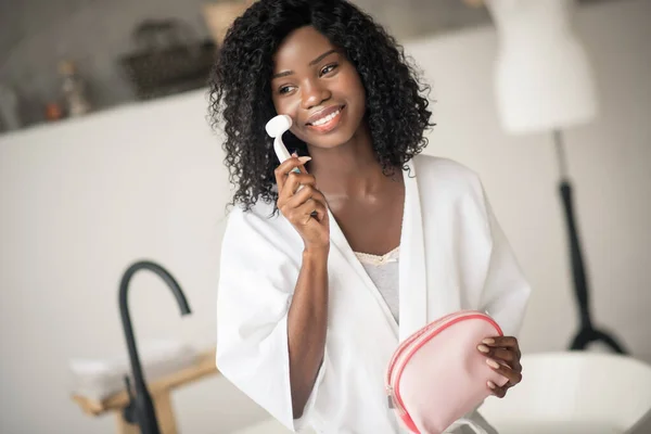 Dark-eyed woman smiling while using facial massage brush — Stock Photo, Image