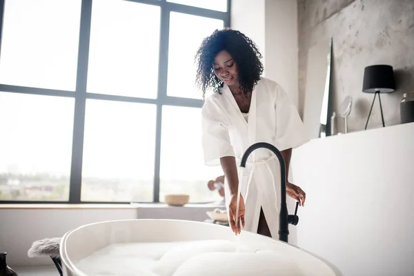 Curly dark-skinned woman preparing bath with foam — Stock Photo, Image