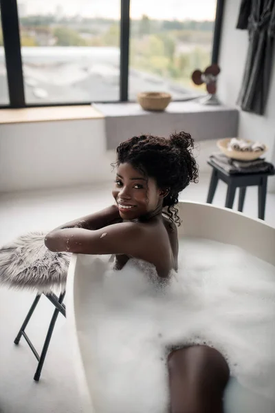 Stralende krullende vrouw gevoel vrolijk terwijl liggend in schuimig bad — Stockfoto