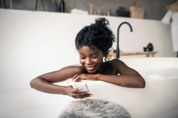 Leende mörkhyade kvinna som ligger i badkaret och använder smartphone — Stockfoto