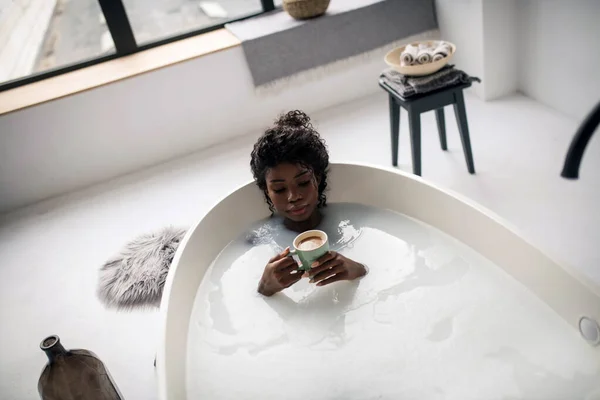 Кудрявая темнокожая женщина пьет капучино, когда лежит в ванной — стоковое фото