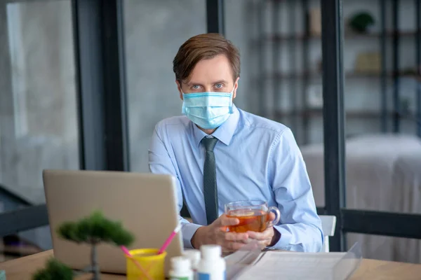 Hombre de negocios enfermo con máscara y beber té caliente en el trabajo — Foto de Stock