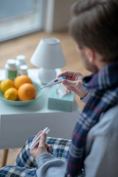 Человек сидит за столом с лекарствами и смотрит на термометр — стоковое фото