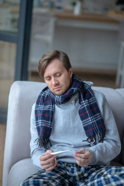 Mann mit Schal sitzt auf Sofa und schaut auf Thermometer — Stockfoto