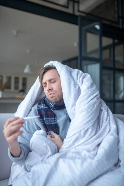Άνδρας καλυμμένος με σεντόνι αισθάνεται λυπημένος μετά τη μέτρηση της θερμοκρασίας — Φωτογραφία Αρχείου