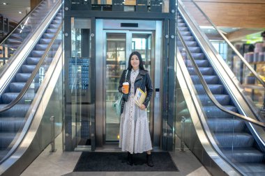 Genç esmer kadın elinde kahve fincanıyla asansörün önünde durmuş gülümsüyor.