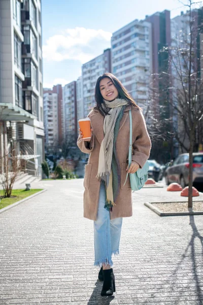 Morena hembra en abrigo beige caminando por la calle con taza de café, sonriendo —  Fotos de Stock