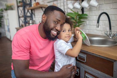 Pembe tişörtlü, uzun boylu, esmer bir adam gülümseyip çocuğuna sarılıyor.