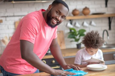 Masayı silip gülümseyen pembe tişörtlü, uzun boylu, esmer bir adam.