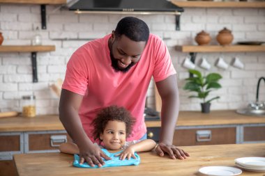 Pembe tişörtlü, uzun boylu, esmer bir adam. Çocuğu otururken ayakta masaları siliyor.