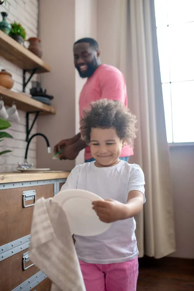 Behoorlijk krullend kind dat de plaat vasthoudt en lacht, haar vader ziet er gelukkig uit. — Stockfoto