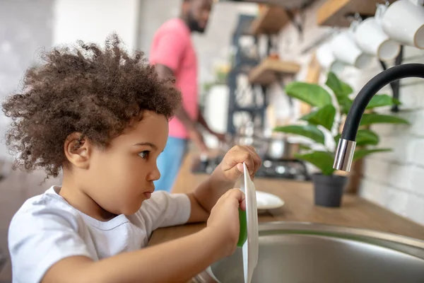 Hübsches lockiges Kind wäscht den Teller und sieht konzentriert aus — Stockfoto