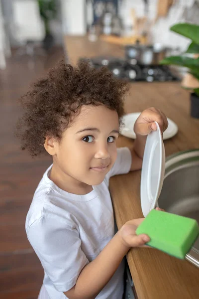 Pretty curly-haired kid holding a plate and a sponge in her hands — Stock Photo, Image