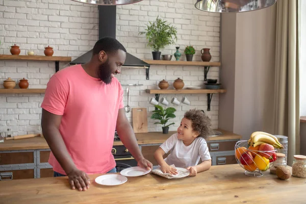 Vysoký tmavovlasý muž v růžové košili stojí a usmívá se, zatímco jeho dítě utírá talíř — Stock fotografie