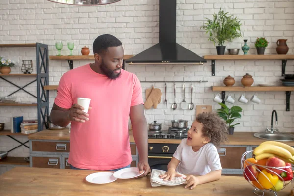Vysoký muž s tmavou pletí v růžovém tričku a jeho kluk utírá talíř — Stock fotografie