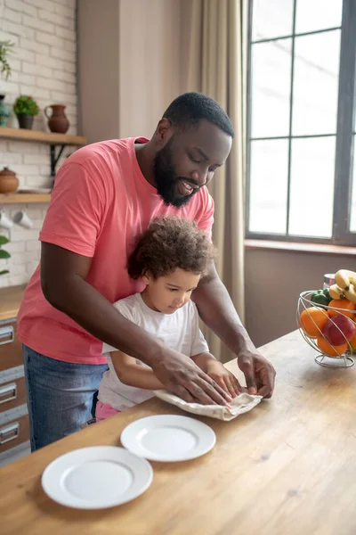 Lange donkere man in een roze tshirt die zijn kind laat zien hoe hij het bord moet afvegen. — Stockfoto