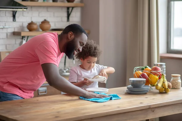 Lang donker gekleurde man in een roze tshirt veegt de tafel af en glimlacht, zijn dochter veegt de plaat af — Stockfoto