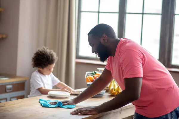 Vysoký tmavovlasý muž v růžovém tričku utírá stůl, zatímco jeho dítě pomáhá — Stock fotografie