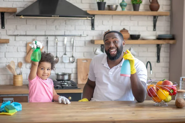 Afro-Amerikaanse bebaarde man en zijn dochter zitten aan tafel met sprays in handen — Stockfoto