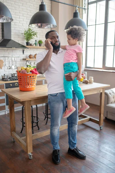 Alto padre dalla pelle scura che sta in piedi e tiene in braccio suo figlio mentre parla al telefono — Foto Stock